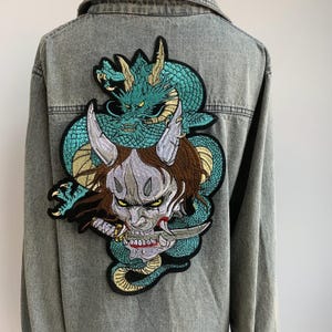 Peut inclure: Veste en jean grise avec un grand écusson brodé au dos. L'écusson représente un dragon turquoise entrelacé avec un masque de démon, avec des cheveux bruns et un couteau. La veste a une fermeture boutonnée et des manches longues.