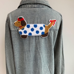 May include: A gray denim jacket with a colorful embroidered dachshund patch on the back. The patch features a brown and white dog with blue polka dots, wearing a red hat and bow.