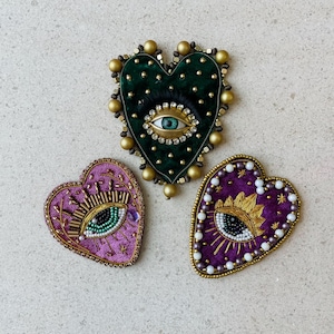 May include: Three heart-shaped brooches featuring an eye design. One is green velvet with gold beads, another is purple with gold and white beads, and the third is purple with gold embroidery and white beads. The brooches are handmade.