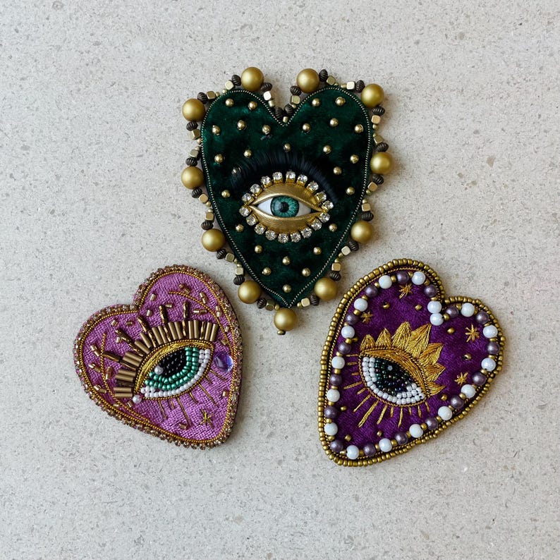 Op de afbeelding: Drie hartvormige broches met een oogontwerp. E&eacute;n is groen fluweel met gouden kralen, een andere is paars met gouden en witte kralen, en de derde is paars met gouden borduursel en witte kralen. De broches zijn handgemaakt.
