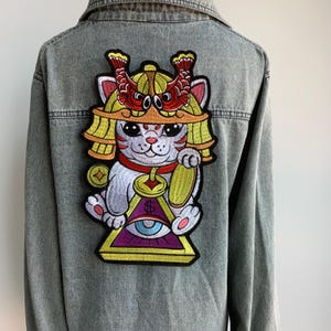 May include: A denim jacket featuring an embroidered patch of a white cat wearing a golden helmet with red accents. The cat is holding a golden bell and sitting on a yellow triangle with an all-seeing eye.