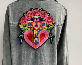 Può includere: Una giacca di jeans con una toppa ricamata colorata che presenta un cuore, una croce e dei fiori. La toppa è un mix vibrante di rosa, rosso, giallo, blu e verde.