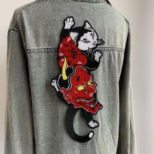 Peut inclure: Une chemise en jean gris d&eacute;lav&eacute; &agrave; l'acide avec un grand &eacute;cusson brod&eacute; dans le dos. L'&eacute;cusson repr&eacute;sente un chat noir et blanc au-dessus d'un masque de d&eacute;mon rouge avec des cornes jaunes et des dents ac&eacute;r&eacute;es, orn&eacute; de motifs floraux rouges.