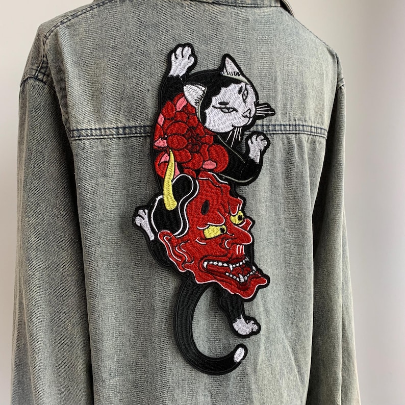 Peut inclure: Une chemise en jean gris d&eacute;lav&eacute; &agrave; l'acide avec un grand &eacute;cusson brod&eacute; dans le dos. L'&eacute;cusson repr&eacute;sente un chat noir et blanc au-dessus d'un masque de d&eacute;mon rouge avec des cornes jaunes et des dents ac&eacute;r&eacute;es, orn&eacute; de motifs floraux rouges.