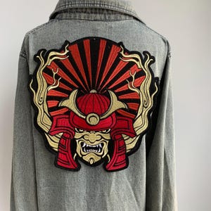 Peut inclure: Veste en jean grise ornée d'un grand écusson brodé dans le dos. L'écusson représente un casque de samouraï rouge et noir avec des détails dorés, sur un motif de soleil rouge et noir. La veste a une fermeture boutonnée et un col.