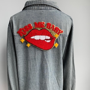 Könnte beinhalten: Graue Jeansjacke mit einem großen, gestickten Aufnäher auf dem Rücken. Der Aufnäher zeigt rote, paillettenbesetzte Lippen mit dem Text "KISS ME BABY" in roter und goldener Schrift. Die Jacke hat eine Knopfleiste und einen Kragen.