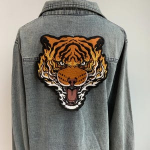 Può includere: Una giacca di jeans con una grande toppa ricamata a forma di testa di tigre sul retro. La tigre è arancione, nera e bianca con un'espressione feroce.