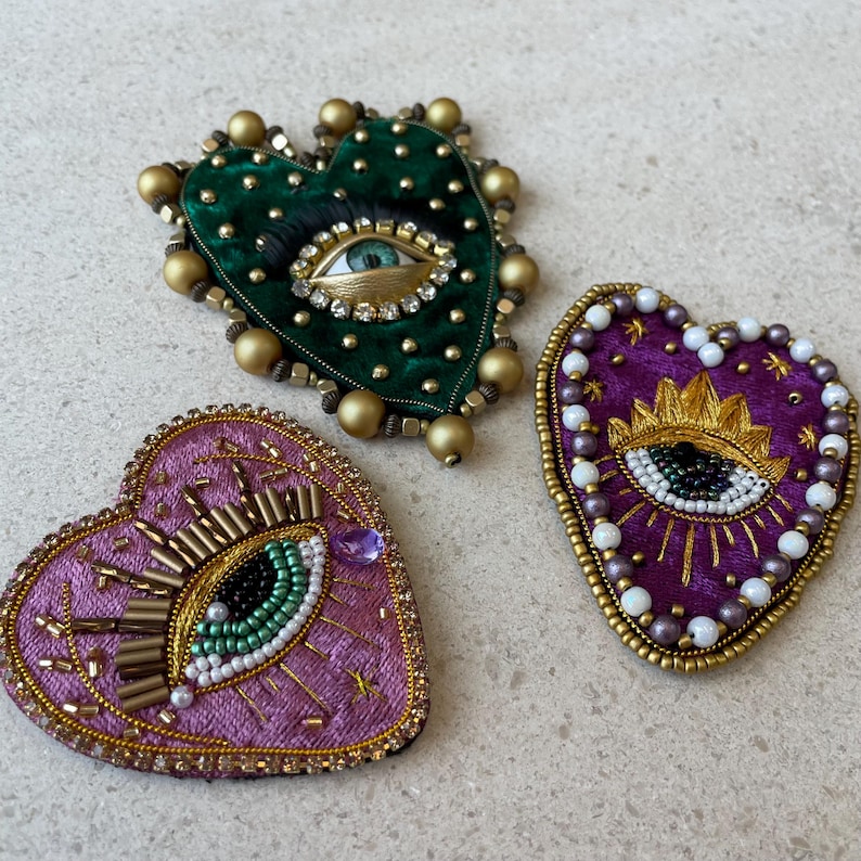 Op de afbeelding: Drie hartvormige broches met een oogontwerp. E&eacute;n is groen fluweel met gouden kralen, een andere is paars met gouden accenten en de derde is paars met gouden en witte kralen. Elke broche heeft een oog in het midden.