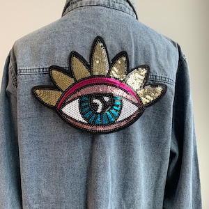 Può includere: Giacca di jeans azzurra con una grande toppa a forma di occhio con paillettes sul retro. La toppa presenta un'iride blu, una palpebra rosa e dettagli dorati e neri. La giacca è abbottonata.