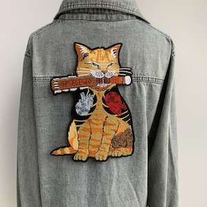 Op de afbeelding: Een grijze spijkerjas met een groot geborduurd embleem van een oranje tijgerkat die een houten voorwerp in zijn bek houdt. De kat heeft ingewikkelde tatoeages en een serene uitdrukking. De jas heeft een knoopsluiting en een kraag.