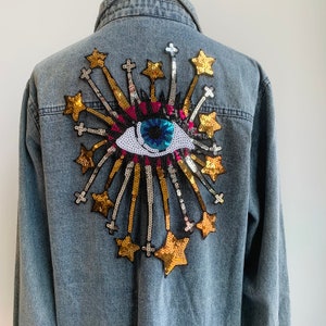 May include: A denim jacket with a large sequined eye and starburst design on the back. The eye is blue, white and pink, and the stars are gold.