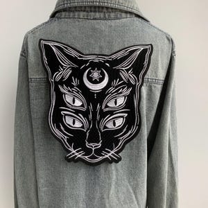 Peut inclure: Patch thermocollant brodé noir représentant un visage de chat avec quatre yeux et une lune croissante au-dessus de sa tête.