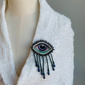 May include: A dark blue eye-shaped brooch with a pink and green iris and a white pearl pupil. The brooch has a fringe of dark blue beads.