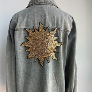 May include: A grey denim jacket featuring a large, circular sun patch on the back. The sun design is crafted from shimmering gold sequins with a black embroidered outline, contrasting against the faded grey fabric of the long-sleeved garment.