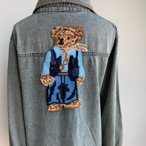 Könnte beinhalten: Eine blaue Jeansjacke mit einem gestickten braunen und blauen Bären, der eine Weste und eine Sonnenbrille auf dem Rücken trägt.