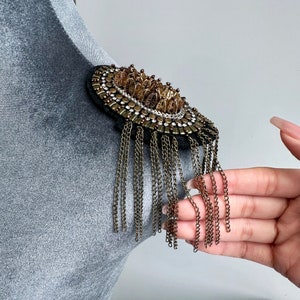 Puede incluir: Un broche circular dorado y marrón con una franja de cadenas. El broche está decorado con cuentas y lentejuelas.