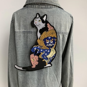 Puede incluir: Una chaqueta vaquera con un parche bordado de un gato blanco y negro y un león azul y dorado.