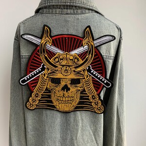 May include: Embroidered patch featuring a golden skull wearing a samurai helmet, crossed swords, and a red sunburst design. The patch is attached to the back of a light-wash denim jacket. The patch is a detailed, decorative element.