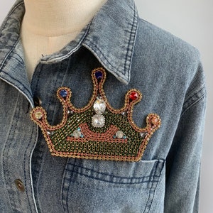 May include: A denim jacket with a beaded crown patch. The crown is green, gold, and red with clear rhinestones.