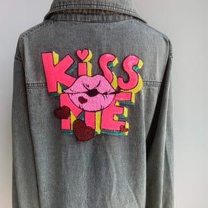 May include: Gray denim jacket with a sequined "Kiss Me" design on the back. The design features pink lips, pink and yellow letters, and red heart accents. The jacket has a classic collar and button-up front.