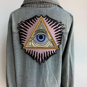 May include: A denim jacket with an embroidered patch of an all-seeing eye inside a triangle. The eye is blue with a white iris and a pink eyelid. The triangle is surrounded by a pink and black sunburst.