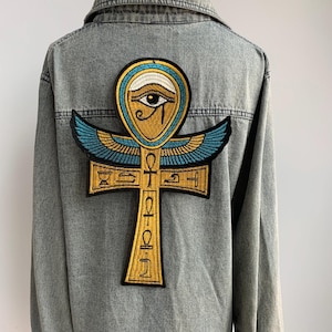 May include: A denim jacket with a large embroidered patch featuring an Egyptian ankh symbol. The patch is gold, blue, and black, with an eye design at the top. The jacket is a light blue wash.