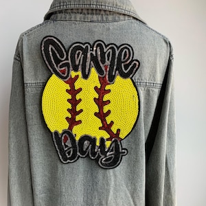 May include: A light gray denim jacket with a large sequined patch on the back. The patch features a yellow softball with red stitching and the words "Game Day" in black sequins.