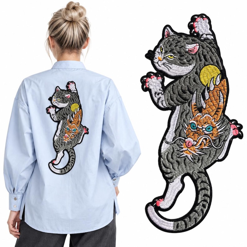 Peut inclure: Une chemise bleu clair &agrave; boutons avec un grand &eacute;cusson brod&eacute; au dos. L'&eacute;cusson repr&eacute;sente un chat gris avec une lune jaune et une cr&eacute;ature orange semblable &agrave; un dragon aux yeux bleus, le tout soulign&eacute; de fil noir avec des d&eacute;tails roses.