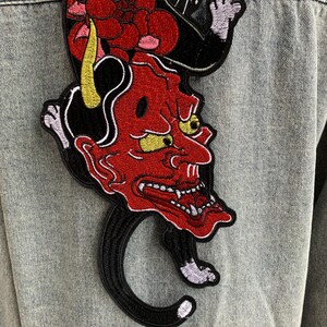 Peut inclure: Une veste en jean bleu clair avec un grand &eacute;cusson brod&eacute; au dos. L'&eacute;cusson repr&eacute;sente un masque de d&eacute;mon au visage rouge avec des cornes jaunes et des dents pointues, plac&eacute; au-dessus d'une silhouette de chat noir avec des motifs floraux rouges.