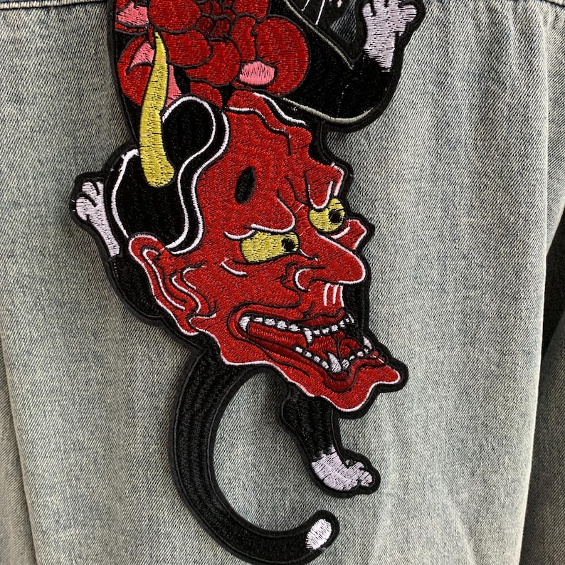Peut inclure: Une veste en jean bleu clair avec un grand &eacute;cusson brod&eacute; au dos. L'&eacute;cusson repr&eacute;sente un masque de d&eacute;mon au visage rouge avec des cornes jaunes et des dents pointues, plac&eacute; au-dessus d'une silhouette de chat noir avec des motifs floraux rouges.