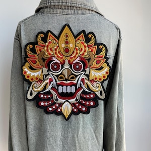 Peut inclure: Veste en jean délavé avec un grand écusson brodé dans le dos. L'écusson représente un masque de Barong balinais, avec des détails rouges, dorés et noirs. Le design de la veste contraste avec le denim.