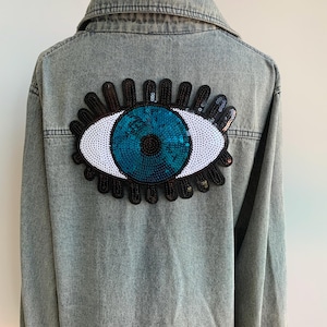 May include: A light-wash denim jacket featuring a large, sequined eye design on the back. The eye has a teal iris, white sclera, and black lashes. The jacket has a classic collar and button-up front.