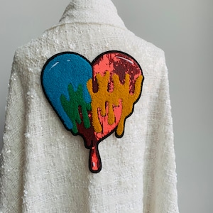Puede incluir: Chaqueta blanca con un parche en forma de corazón de colores. El parche está hecho de lentejuelas y tiene un efecto de goteo. Los colores del parche son rojo, azul, verde y amarillo.
