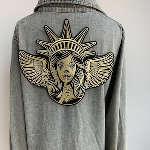 May include: A gray denim jacket with a large embroidered patch on the back. The patch features a gold-colored design of a woman with a crown, wings, and a finger to her lips, in a 'shhh' gesture.