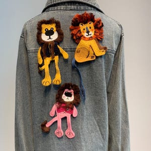 Puede incluir: Una chaqueta vaquera con tres parches bordados de leones. Los leones son amarillos, naranja y rosa. Los parches están cosidos en la parte posterior de la chaqueta.