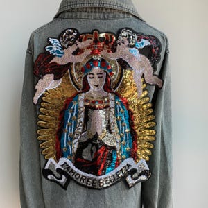 Può includere: Una giacca di jeans con una grande toppa di paillettes colorate sul retro. La toppa presenta una figura religiosa con angeli e le parole "L'AMORE E' BELLEZZA".