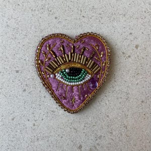 Op de afbeelding: Een hartvormige, geborduurde patch in paars, goud en groen. De patch heeft een oogontwerp met gouden kralen, een groene iris en een paarse edelsteen. De randen zijn afgewerkt met gouden kralen.