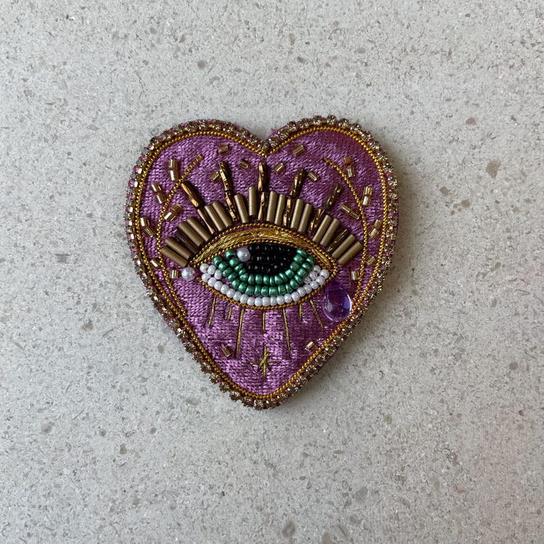 Op de afbeelding: Een hartvormige, geborduurde patch in paars, goud en groen. De patch heeft een oogontwerp met gouden kralen, een groene iris en een paarse edelsteen. De randen zijn afgewerkt met gouden kralen.