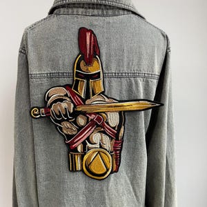 Puede incluir: Chaqueta vaquera gris claro con un gran parche bordado en la espalda. El parche representa a un guerrero espartano dorado y rojo sosteniendo una espada. El guerrero lleva un casco y tiene un escudo en la parte inferior.