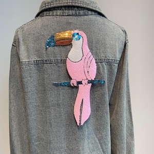 Może przedstawiać: Dżinsowa kurtka z haftowanym różowym i złotym tukankiem na plecach. Tukan siedzi na gałęzi.