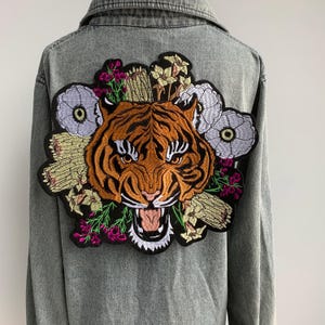 Op de afbeelding: Grijze spijkerjas met een grote geborduurde tijgerkop omringd door kleurrijke bloemmotieven. De tijger is oranje en zwart, met witte tanden en een zwarte omtrek. De bloemen hebben paarse, gele en witte details.