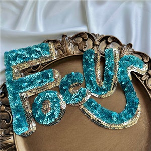 Puede incluir: La palabra "Forever" escrita en lentejuelas turquesa y doradas. Las lentejuelas están dispuestas en forma de onda.