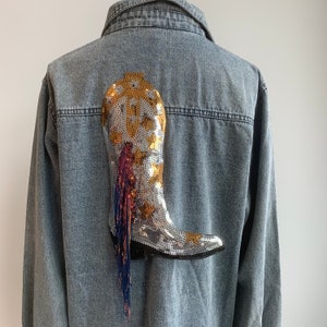 May include: A denim jacket with a silver sequined boot appliqué. The boot is decorated with gold and red sequins and has a fringe detail.