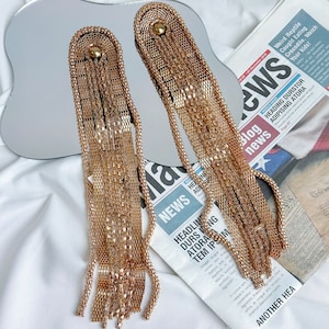 Puede incluir: Un par de pendientes de cadena dorada con borlas. Los pendientes son largos y tienen un diseño delicado de cadena. Las borlas están hechas de múltiples hebras de cadena que están conectadas a la cadena principal.