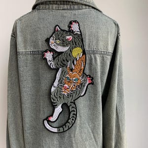 Op de afbeelding: Een grijze spijkerjas met een grote geborduurde patch op de rug. De patch toont een gestileerd kattenontwerp met een draak verwerkt in zijn lichaam, in tinten grijs, oranje, geel en wit. De jas heeft een klassieke kraag en een knoopsluiting.
