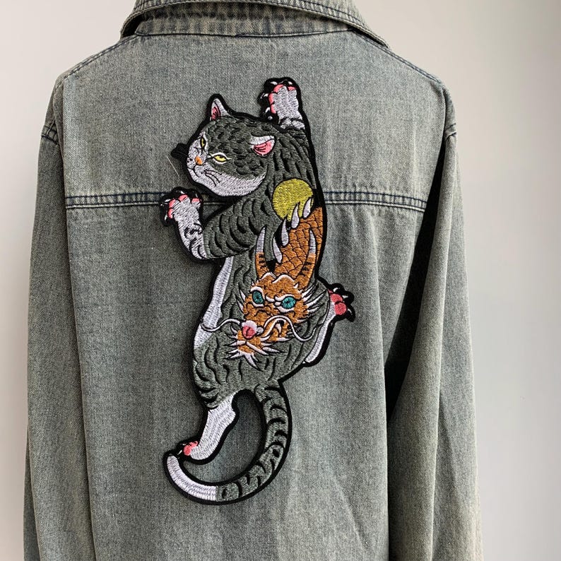 Peut inclure: Une veste en jean gris avec un grand &eacute;cusson brod&eacute; au dos. L'&eacute;cusson repr&eacute;sente un chat tigr&eacute; gris avec une lune jaune au-dessus d'une cr&eacute;ature orange plus petite ressemblant &agrave; un dragon, aux yeux bleu sarcelle et moustaches blanches.