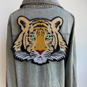 Può includere: Giacca di jeans grigio chiaro con una grande toppa ricamata a forma di tigre sul retro. La toppa della tigre presenta dettagli dorati, neri e bianchi, con occhi verdi e naso rosso. La giacca ha un colletto classico.