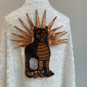 May include: A white jacket with a brown and black embroidered cat with a gold sequin halo. The cat is sitting with its tail curled around its legs.