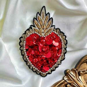 Può includere: Un broche a forma di cuore rosso con decorazioni dorate e argentate. Il cuore è ricoperto di paillettes rosse e circondato da un bordo di perline.