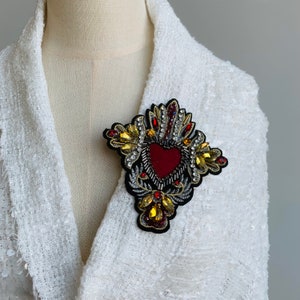 Puede incluir: Un broche en forma de corazón rojo con adornos dorados y plateados. El corazón está rodeado de un diseño decorativo de flores y hojas.
